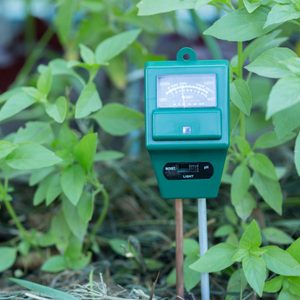 A green soil moisture meter stands among green plant leaves, measuring soil moisture levels in a garden setting.