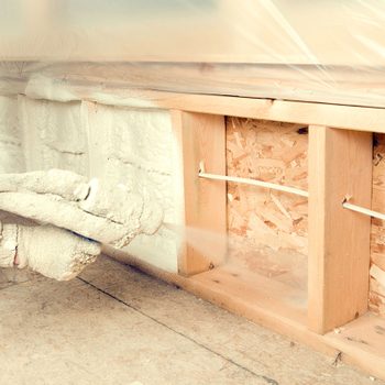 A person applies spray foam insulation to wall cavities in a construction area, surrounded by wooden frames and plastic sheeting to contain the mess.