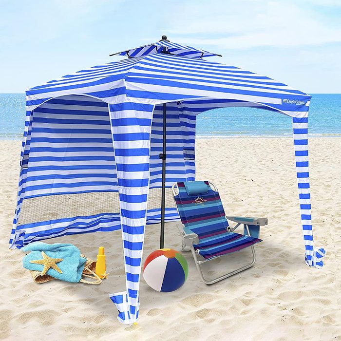 A blue and white striped beach tent provides shade. A striped chair sits nearby, alongside a beach ball and sunscreen, with the ocean in the background.