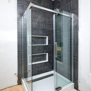 A glass shower enclosure stands against a patterned black wall, featuring a sleek chrome handle and a white base, creating a modern bathroom atmosphere.