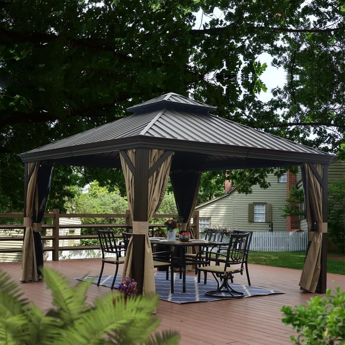 A gazebo stands on a wooden deck, featuring a table and chairs underneath. Surrounding trees and a house create a serene outdoor setting.