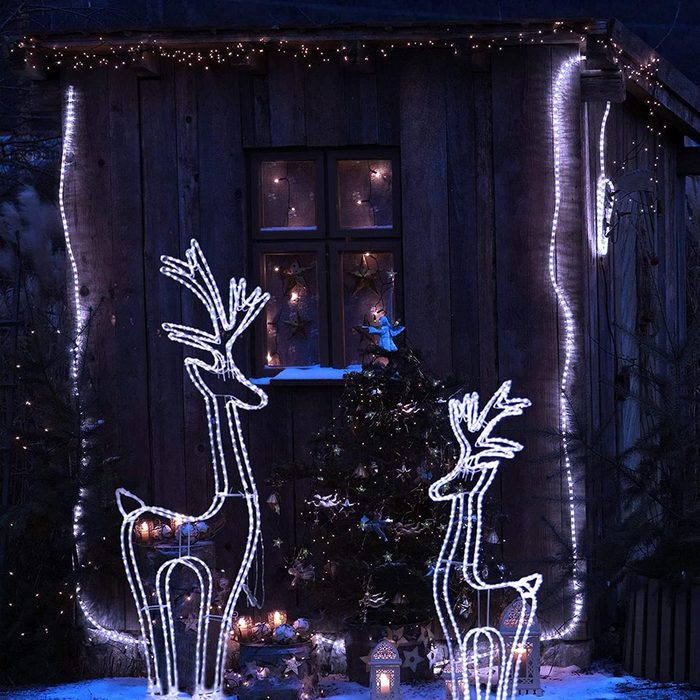 Two illuminated reindeer decorations stand in snow, while a cozy wooden shed is adorned with twinkling lights, surrounded by festive trees and holiday decorations.
