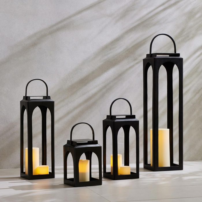 Four black lanterns with illuminated candles are arranged together, casting shadows on a light gray wall in a minimalist setting.