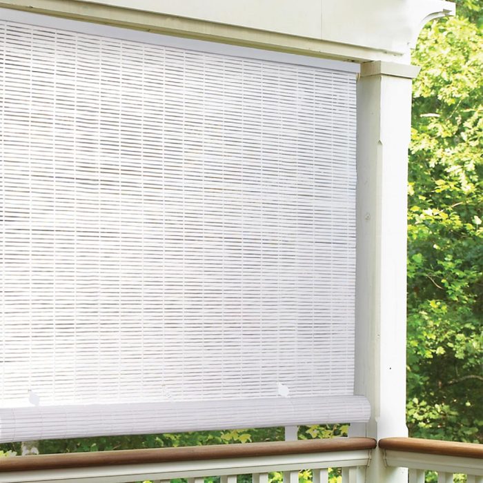 A white bamboo roller shade covers a porch railing, filtering sunlight while greenery is visible in the background, creating a tranquil outdoor atmosphere.