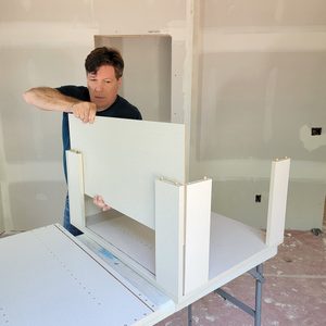 A man positions a flat panel onto a partially assembled wooden structure on a table in a room with unfinished drywall.