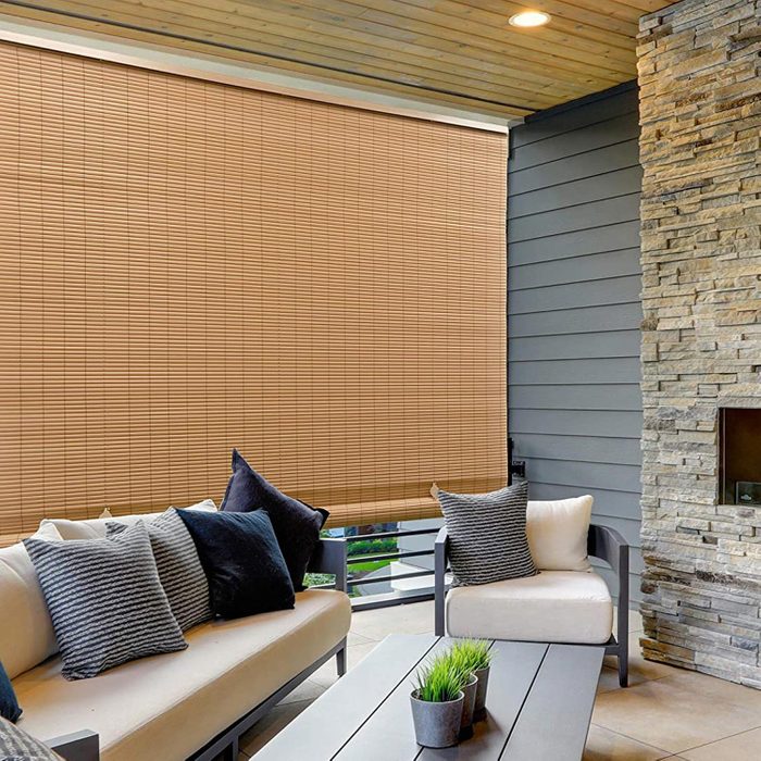 A cozy outdoor seating area features a light-colored couch adorned with various cushions. Bamboo blinds cover a wall, surrounded by stone and wood accents.