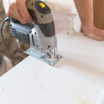 A person operates a jigsaw, cutting through a white surface, with wood shavings visible. The background features various items, suggesting a workshop environment.