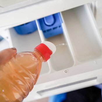 A hand holds a bottle with a red cap, pouring liquid into an empty compartment inside a white appliance, with colorful items visible in the background.
