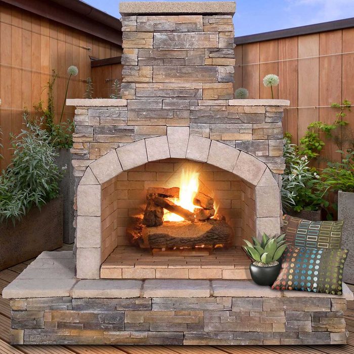 A stone fireplace burns logs, radiating warmth. Surrounding greenery and decorative pillows create a cozy outdoor atmosphere against a wooden backdrop.