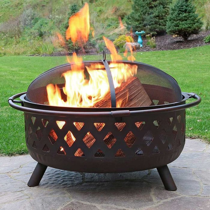 A fire pit with flames roaring inside burns logs, set on a stone patio amid a green lawn and trees in the background.