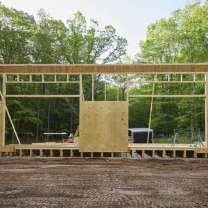 A wooden frame structure stands incomplete with a large wooden panel. It's situated in a lush, green forested area, with construction tools in the background.
