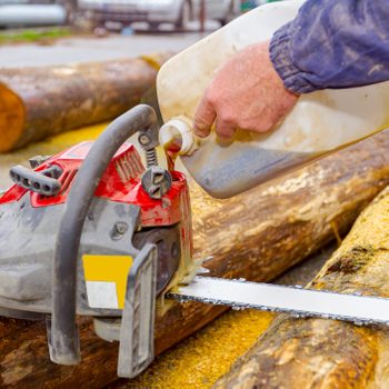 A person pours oil into a chainsaw, preparing it for use, with logs and sawdust scattered around in a busy outdoor environment.