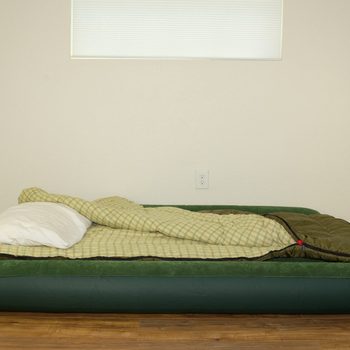An inflatable mattress rests on wooden flooring, covered with a plaid blanket and a white pillow, in a minimally decorated room with a small window.