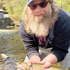 A man with a beard holds a colorful fish in shallow water, surrounded by greenery and a wooden structure in the background.