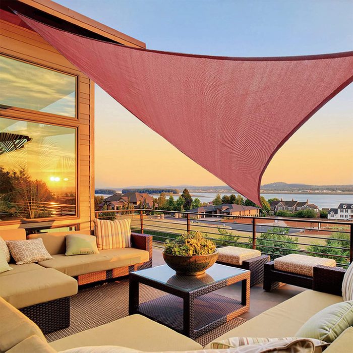 A large, triangular shade sails above a stylish outdoor seating area, with a sunset casting warm light over a scenic view of houses and water.