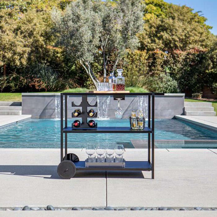 A black bar cart holds liquor bottles and glasses next to a serene pool, framed by lush greenery and a modern water feature.