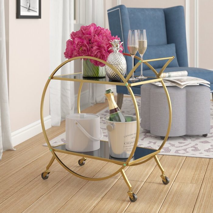 A gold bar cart holds a bouquet of pink flowers, a pineapple, glasses of champagne, and a bottle of sparkling wine, set in a cozy room.