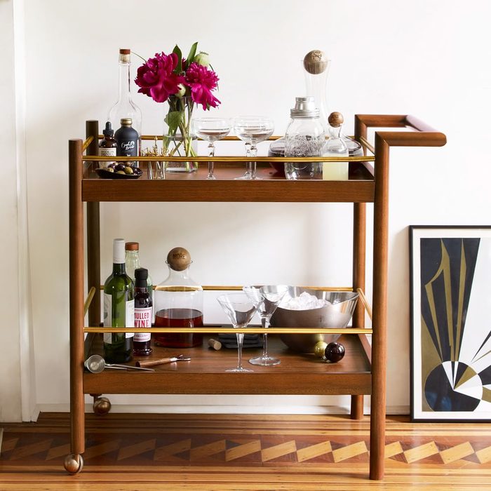 A wooden bar cart displays wine bottles, glassware, and a bouquet of flowers, set against a minimalist wall and geometric artwork.