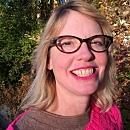 A smiling woman with blonde hair and glasses stands outdoors, surrounded by greenery and autumn leaves. She appears cheerful and engaged with her surroundings.