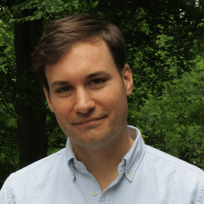 A young man with short brown hair stands smiling in front of a backdrop of green foliage, wearing a light blue button-up shirt.