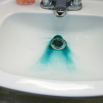 A white sink contains water with a blue-green discoloration surrounding the drain, suggesting a possible cleaning agent or dye. Soap is nearby.
