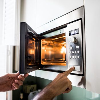 A hand presses a button on an open microwave, which is illuminated inside, set in a kitchen with natural light from a nearby window.