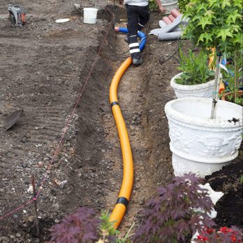 An orange pipe is laid along a trench, while a worker moves nearby, surrounded by potted plants and gardening tools in a landscaped area.