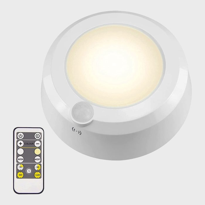 A round, white LED lamp emits soft light, while a remote control with buttons for settings and timer options rests beside it, set against a plain background.
