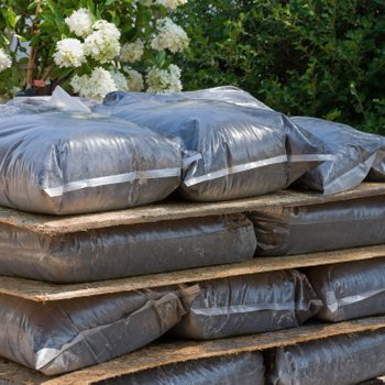 Plastic bags filled with dark material are stacked on wooden pallets, surrounded by greenery and white flowering plants in a bright outdoor setting.
