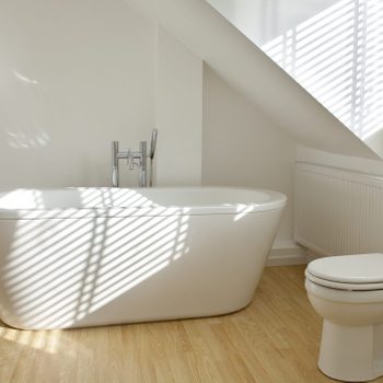 A freestanding bathtub is illuminated by sunlight filtering through blinds, positioned beside a toilet and a radiator on a wooden floor in a minimalist bathroom.