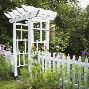 A white garden arbor stands surrounded by colorful flowers and a picket fence, set against a lush green backdrop of trees.