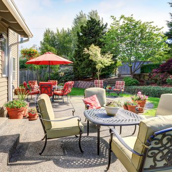 A round table with chairs sits on a patio. Potted plants surround it, while bright umbrellas shade distant seating in a vibrant garden.