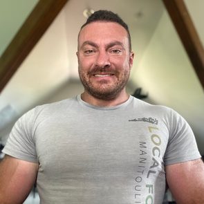 A man with a beard smiles while wearing a grey shirt. He is in a well-lit indoor space with gym equipment visible in the background.