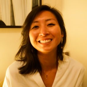 A woman smiles brightly while sitting indoors, illuminated by soft light, with curtains and a mirror visible in the background.