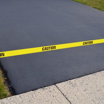 A yellow caution tape lines a fresh, dark asphalt surface, which is bordered by green grass and adjacent to a light-colored concrete path.
