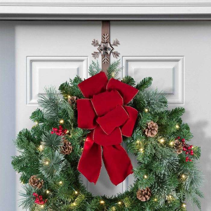A decorative wreath adorned with red ribbon and pinecones hangs on a white door, surrounded by green foliage and small lights.