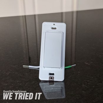 A white electrical switch rests on a dark, smooth surface, displaying wires attached to the bottom, under soft lighting from an overhead source.