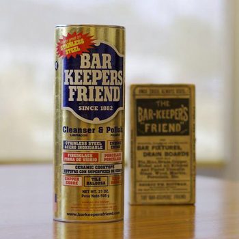 A can of Bar Keepers Friend cleaner stands beside an old, vintage box of the same product, on a wooden surface with natural light in the background.