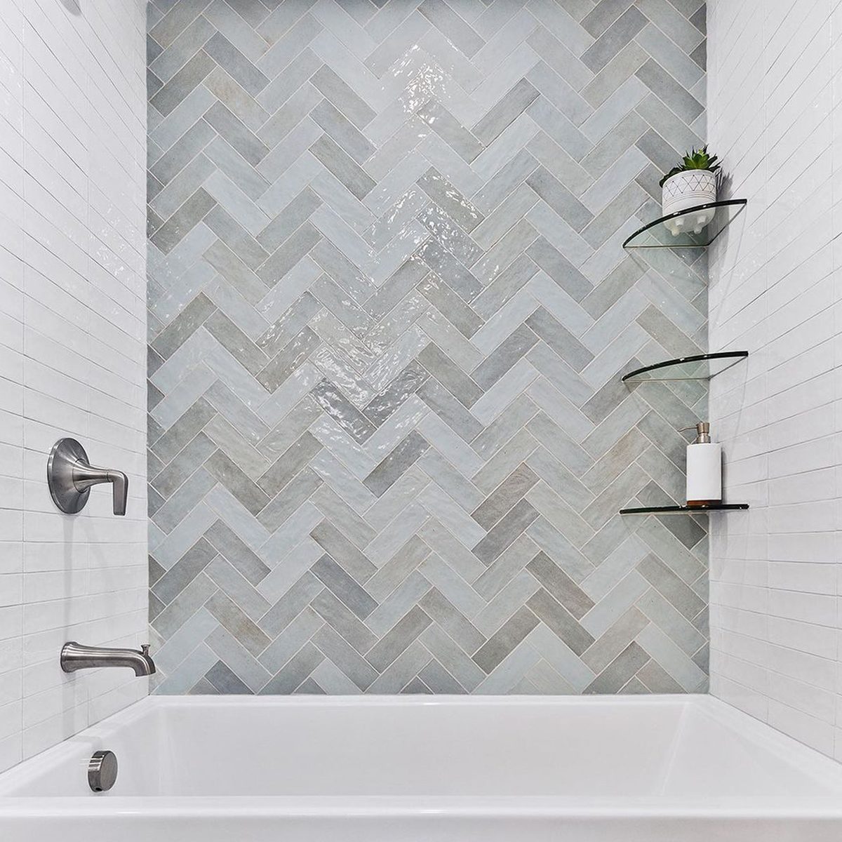 A white bathtub sits against a stylish herringbone-patterned tile wall, featuring gray and blue shades, with glass shelves holding decorative items.