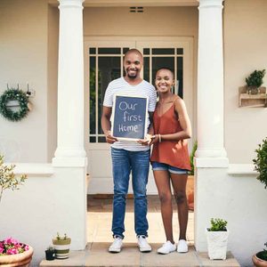 A couple stands together on a porch holding a sign that reads 
