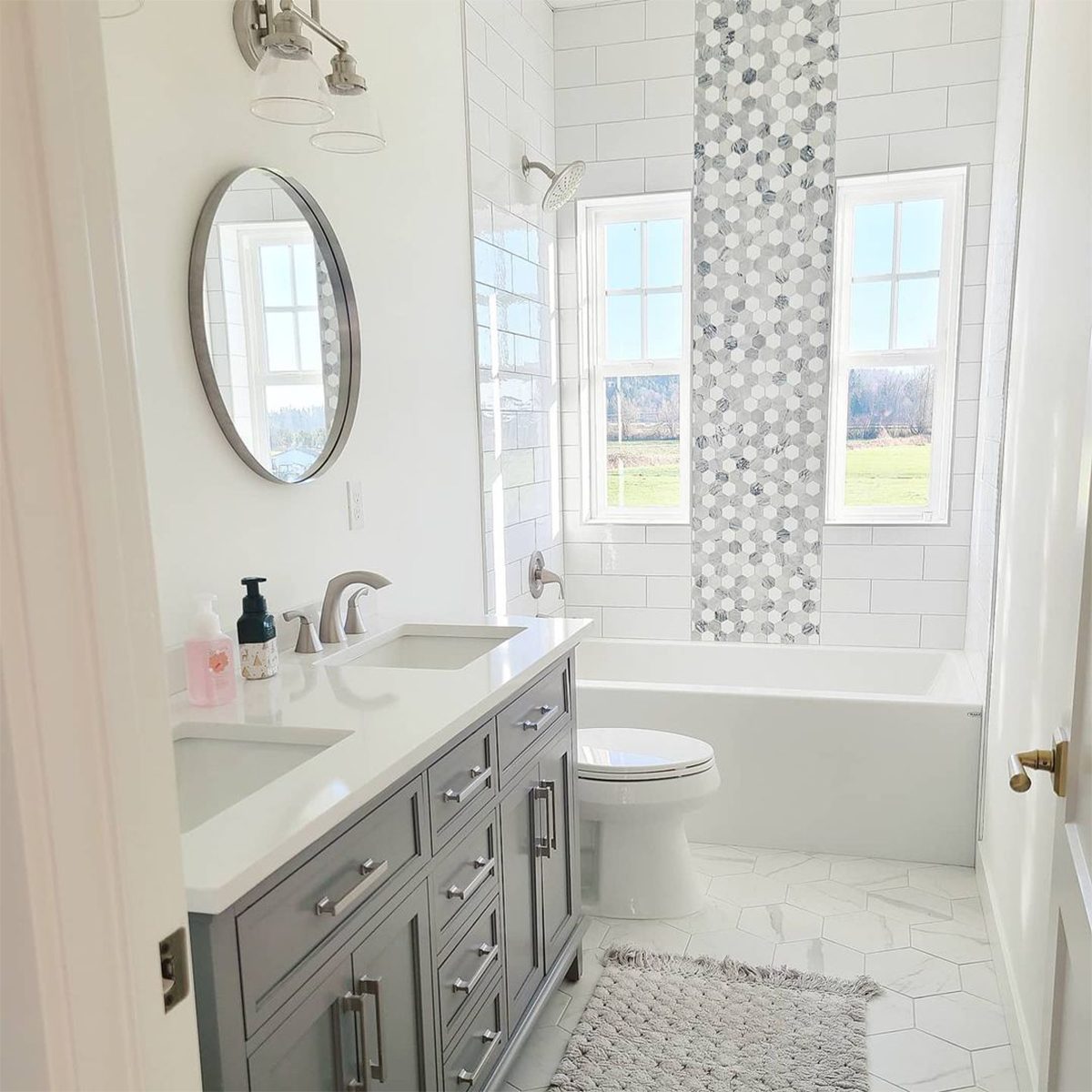 A grey bathroom vanity with sinks reflects light, while a shower head and tub await use near large windows overlooking a grassy landscape.