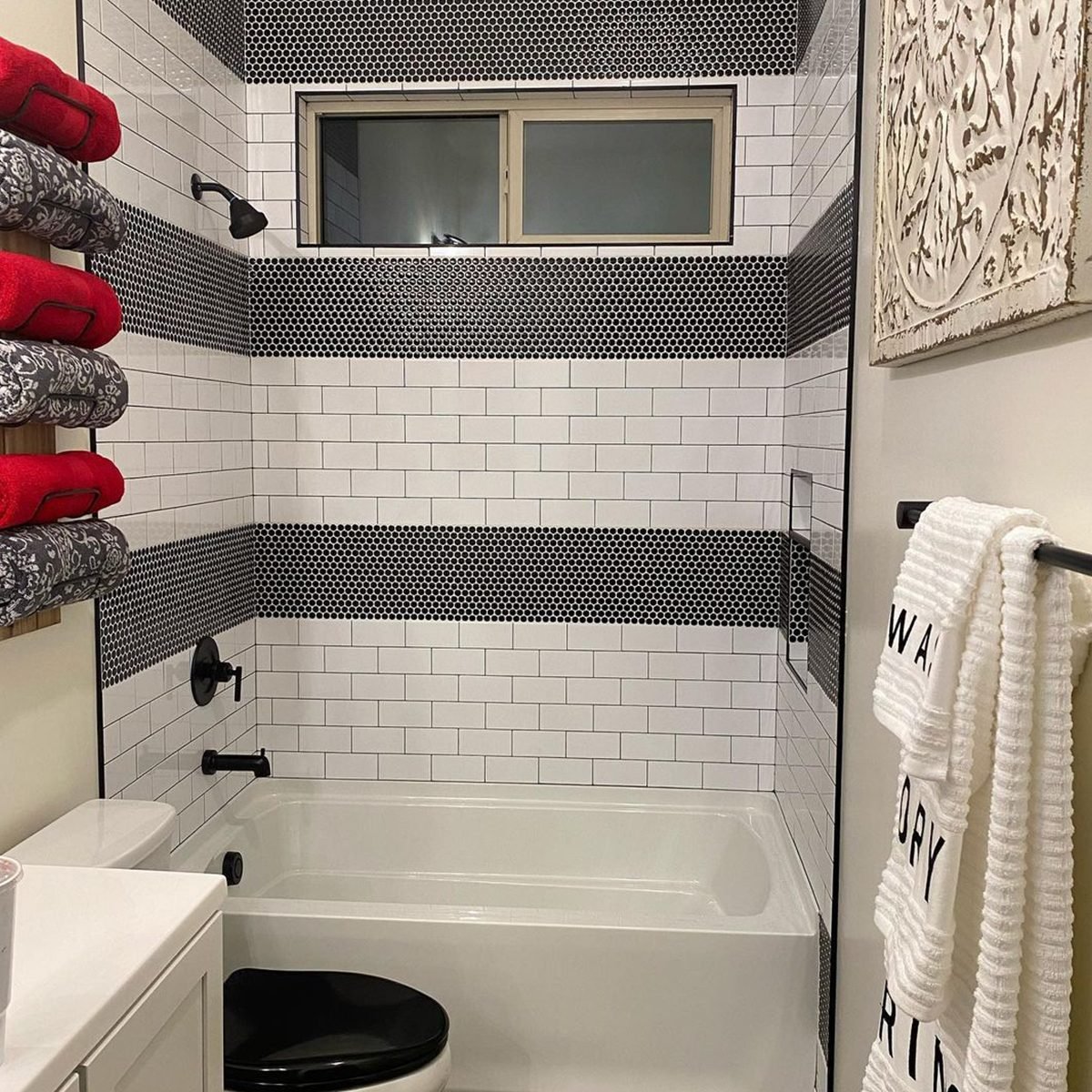 A bathroom features a white bathtub, black fixtures, and a window. Red towels are stacked on one wall, while a patterned towel hangs nearby.