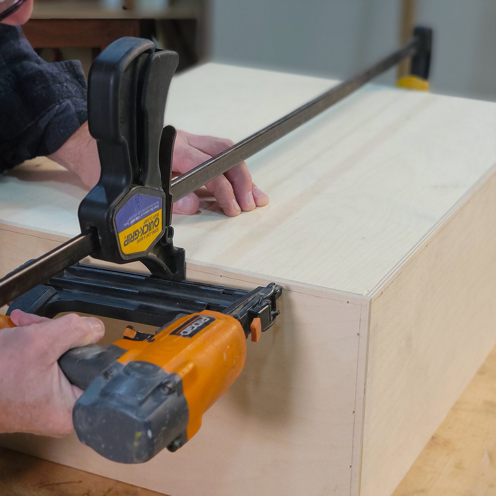 A person uses a nail gun to secure a wooden panel, while a clamp holds the piece steady in a workshop environment.