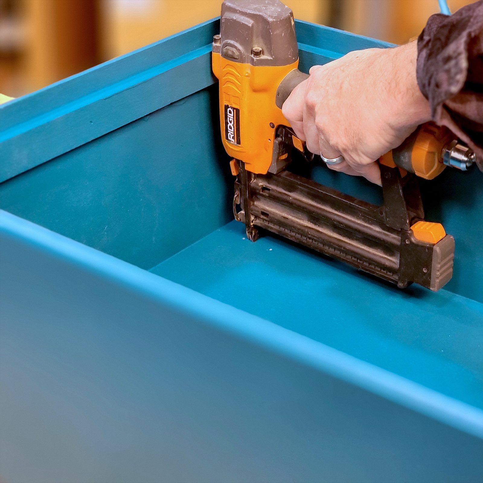 A person uses a cordless nail gun to secure material inside a blue box in a workshop setting, surrounded by tools and light wood surfaces.