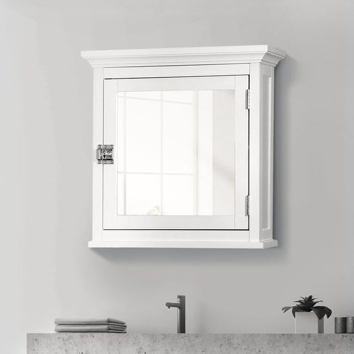 A rectangular white medicine cabinet with a mirror is mounted on a wall, reflecting light, above a gray countertop with folded towels and a small plant.