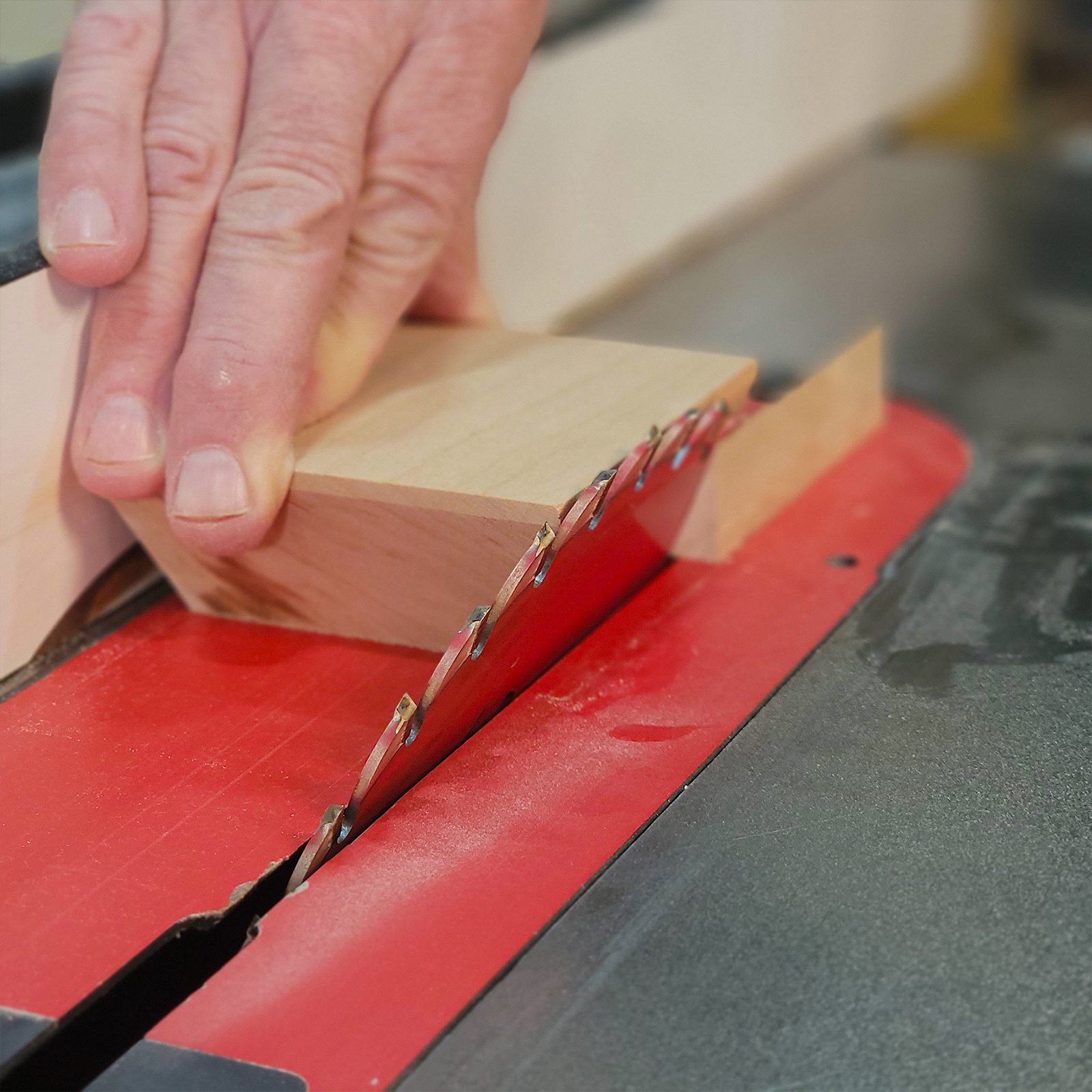 A hand presses a wooden block against a circular saw blade, cutting it on a tabletop surface in a workshop environment.
