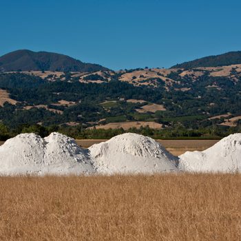Three large white mounds of material are situated in a dry, golden field, with green hills and a clear blue sky in the background.