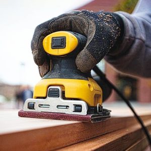 A hand holds a yellow electric sander, smoothing a wooden surface outdoors, with blurred figures in the background and a building behind.