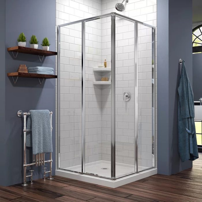 A glass shower stall stands in a modern bathroom, with towels on a rack nearby and plants on shelves against a tiled wall.