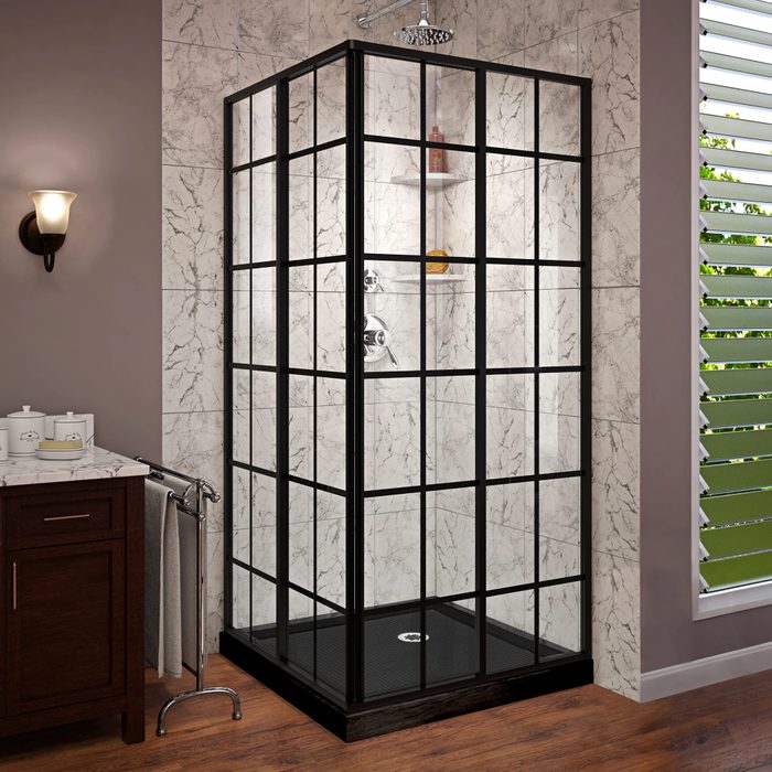 A glass shower enclosure stands in a bathroom, featuring a black frame, fixtures inside, with a wooden cabinet and a lit sconce nearby.